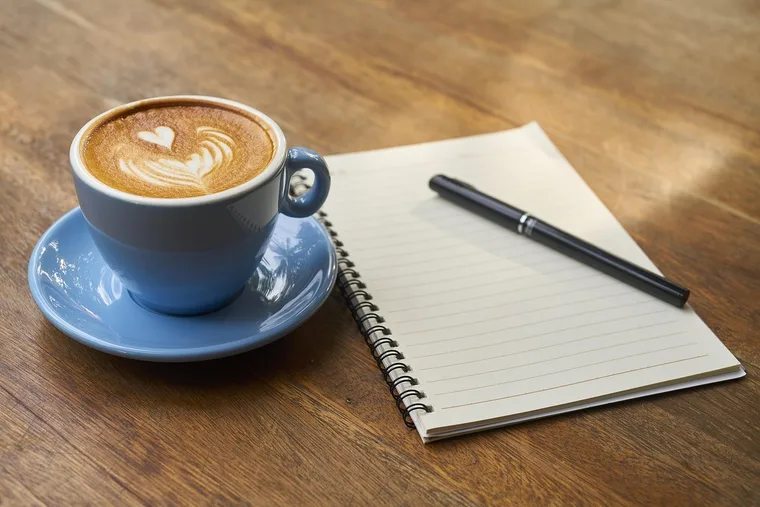 Xícara de café ao lado de um bloco de anotações e uma caneta, sobre mesa de madeira